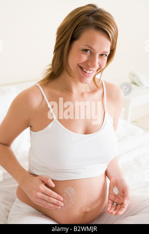 Donna incinta nella camera da letto di crema di sfregamento sulla pancia sorridente Foto Stock