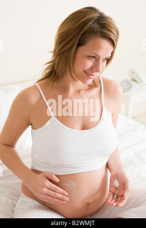 Donna incinta nella camera da letto di crema di sfregamento sulla pancia sorridente Foto Stock