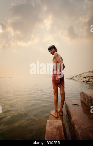 Un giovane ragazzo indiano sorge vicino al bordo di un fiume e si guarda indietro alla videocamera prima di immersioni a. Foto Stock