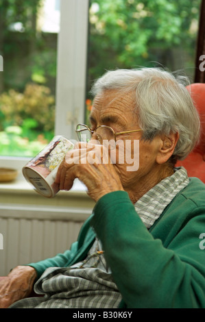 Donna anziana di 92 anni seduta bere il tè da una tazza in salotto Foto Stock