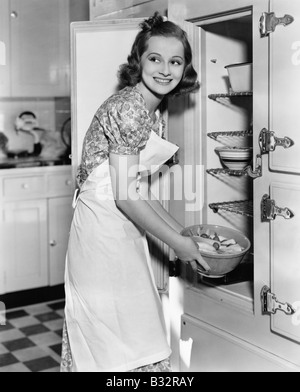 Giovane donna in un grembiule nella sua cucina prendendo il cibo fuori dal frigorifero Foto Stock