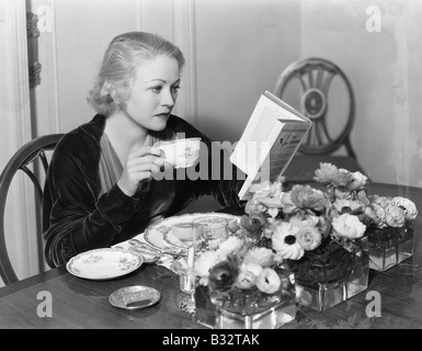 Giovane donna seduta al tavolo da pranzo tenendo una tazza di tè e la lettura di un libro Foto Stock