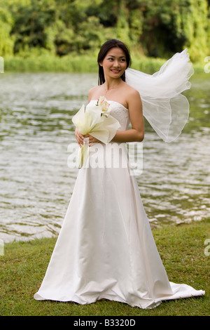 Una donna asiatica si erge nei pressi di un lago nel suo abito da sposa il velo battenti nel vento Foto Stock