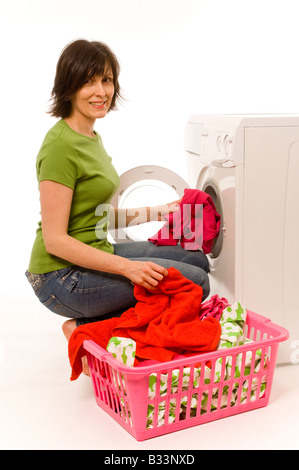 Giovane donna caucasica che indossa una t-shirt verde e jeans che caricano la lavatrice domestica con abiti dai colori vivaci dal cesto della lavanderia. Foto Stock