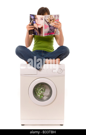 Femmina caucasica con capelli scuri che indossa una parte superiore verde e jeans blu, seduto a croce sulla parte superiore di una lavatrice domestica che legge una rivista. Foto Stock