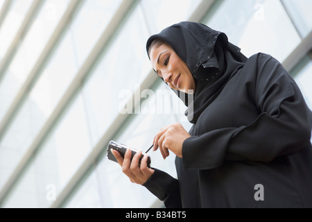 Imprenditrice all'aperto da edificio utilizzando assistente digitale personale e sorridente (messa a fuoco selettiva) Foto Stock