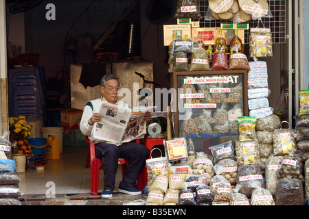 Venditore leggendo un giornale nel suo negozio nella città vecchia, Hanoi, Vietnam Foto Stock