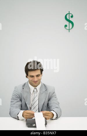 Ragioniere mediante aggiunta di una macchina, sorridente, il simbolo del dollaro sulla parete dietro di lui Foto Stock