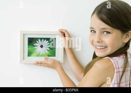 Bambina azienda foto incorniciate contro la parete, sorridente sulla spalla in telecamera Foto Stock