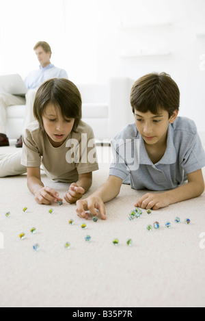 Ragazzi sdraiato sul pavimento a giocare con marmi, padre seduto sul divano con il computer portatile in background Foto Stock