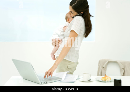 Professional donna in piedi accanto al tavolo per la colazione, tenendo baby, mediante telefono cellulare e computer laptop Foto Stock