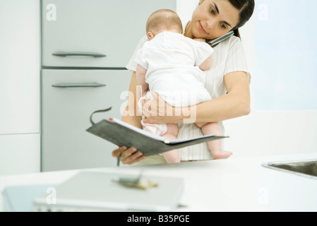 Donna che mantiene baby, tramite telefono cellulare e alla ricerca in agenda Foto Stock