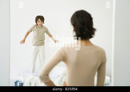 Ragazzo di saltare sul letto, madre in piedi nella porta con la mano sulla sua anca Foto Stock