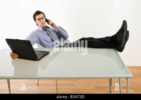 Imprenditore asiatico seduto alla scrivania parlando al cellulare e utilizzando computer portatile Foto Stock