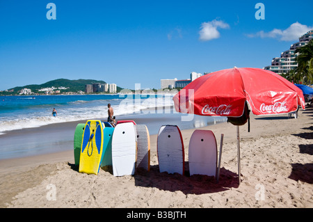 Palmar Bay Beach Ixtapa Mexico / IXTAPA, Guerrero, Messico - Una vivace scena di spiaggia si svolge a Palmar Bay, con un ombrello rosso Coca-Cola e diversi tavolini disposti sulla sabbia. Le persone apprezzano le acque cristalline e le onde dolci, mentre gli edifici del resort fiancheggiano la costa lontana sotto un cielo luminoso. Foto Stock