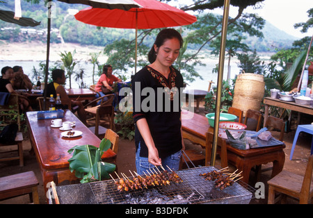 Una donna barbecue spiedini di carne ad un ristorante all'aperto situato sulle rive del fiume Mekong a Luang Prabang, Laos. Foto Stock