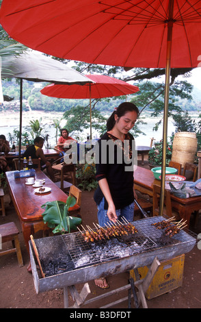 Una donna barbecue spiedini di carne ad un ristorante all'aperto situato sulle rive del fiume Mekong a Luang Prabang, Laos. Foto Stock