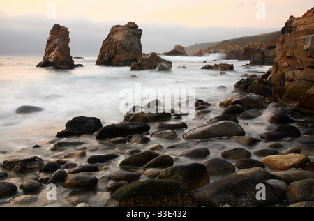 Una spiaggia rocciosa con le onde in movimento con due pinnacoli che fuoriescono dall'acqua in background. Foto Stock