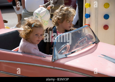 Barneville, Normandia, Francia. Due ragazze in un modello rosa Cadillac su un allegro andare round (giostra) nel mercato settimanale Foto Stock