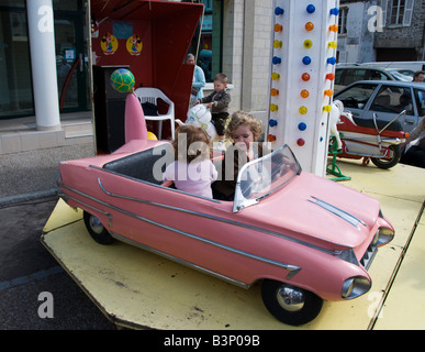 Barneville, Normandia, Francia. Due ragazze in un modello rosa Cadillac su un allegro andare round (giostra) nel mercato settimanale Foto Stock