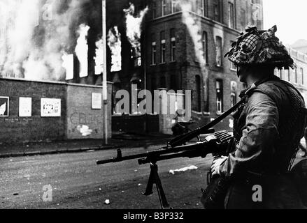Disordini a Belfast le truppe britanniche di guardia a Falls Road Belfast soldato in piedi con una pistola guardando un edificio in fiamme Foto Stock