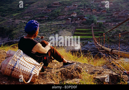 La Cina, nella provincia dello Yunnan, Fiume Yangtse. Il fumo il suo tubo, una donna Naxi riposa dopo aver attraversato un ponte tubo sopra il Yangtse Foto Stock