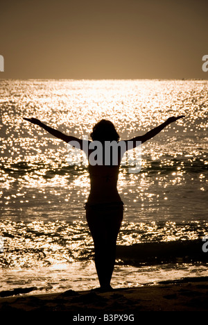 Modello rilasciato donna con le braccia tese stagliano contro il mare splendente dal sole Foto Stock