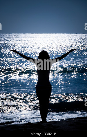 Modello rilasciato donna con le braccia tese stagliano contro il mare splendente dal sole Foto Stock