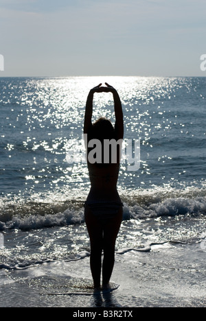 Modello rilasciato donna con le braccia tese stagliano contro il mare splendente dal sole Foto Stock