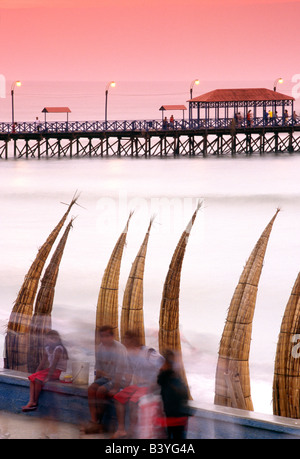 Il Perù, La Libertad, Huanchaco. Totora (reed) barche sono impilati lungo la spiaggia, con il molo di legno in background al villaggio di pescatori di Huanchaco, vicino a Trujillo nel nord del Perù. Foto Stock