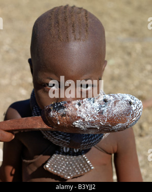 La Namibia, il Kaokoland. Una ragazza Himba bevande latte da un cucchiaio di legno. La sua grande, girocollo, costellata di perle bianche, è chiamato Foto Stock