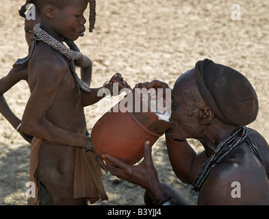 La Namibia, il Kaokoland. Un vecchio uomo Himba bevande latte proveniente da un recipiente di legno trattenuto da una delle sue figlie. Gli Himba sono Herero-spea Foto Stock