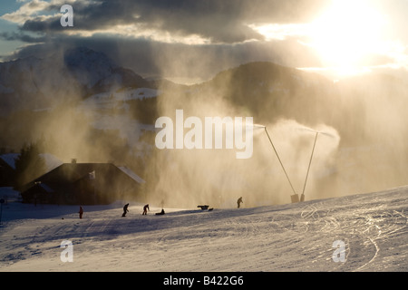 Una nuvola d oro di neve artificiale da impianti di innevamento programmato nelle Alpi francesi, sopra gli sciatori su un pendio nel sole di setting. Foto Stock