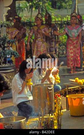 Giovani donne Thai inginocchiato in preghiera con Tailandese classica ballerini eseguono dietro al Santuario di Erawan, Centro città, Bangkok, Thailandia Foto Stock