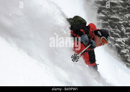 Uno sciatore facendo un giro nella neve profonda nella parte anteriore di un cavalletto di alberi e lasciando un grande spruzzo di neve dietro Foto Stock