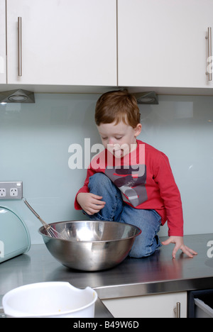 Ragazzo di tre anni e mezzo seduta sul banco di cucina desiderosi di leccare la miscela per torte ciotola Foto Stock