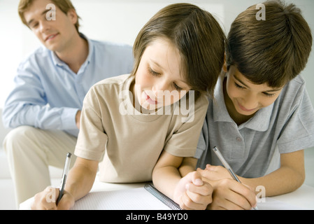 Due ragazzi la scrittura in notebook insieme, padre guardando in background Foto Stock