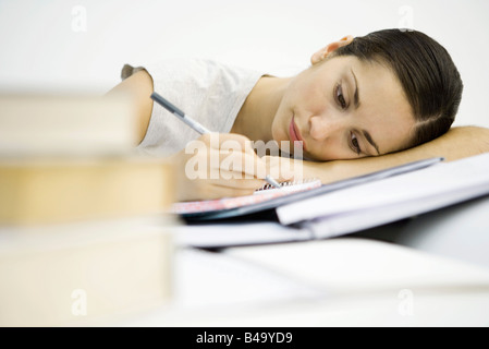 Giovane donna con pile di libri, iscritto nel notebook, testa in appoggio su bracci Foto Stock