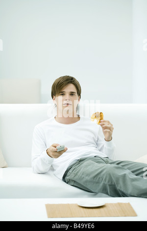 Giovane uomo seduto sul divano, tenendo in mano il telecomando e croissant Foto Stock