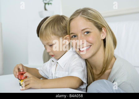 Madre appoggiata la testa sulla spalla del figlio, sorridente in telecamera, ragazzo giocando con automobili giocattolo Foto Stock