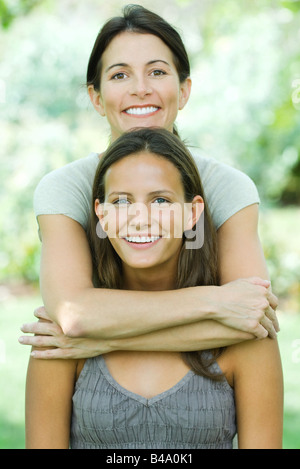 Madre in piedi dietro la figlia adolescente, abbracciando il suo, sia sorridente in telecamera, ritratto Foto Stock