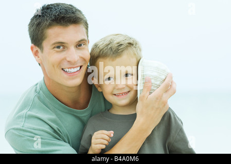 Padre di conchiglia di contenimento al figlio l orecchio, entrambi sorridendo, ritratto Foto Stock