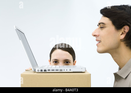 Uomo che utilizza il computer portatile in una scatola di cartone scrivania, donna nascondere dietro la scatola, spiata sopra il bordo Foto Stock