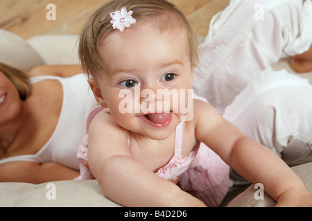 Giovane madre giacente su un divano con i suoi nove mesi di età bambina in piedi di raggiungere con la sua lingua fuori tirando una faccia Foto Stock