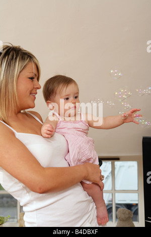 Un felice giovane madre tenendo la sua bambina tra le sue braccia mentre lei gioca con le bolle di sapone. Foto Stock