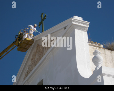 Un pittore su un cherry picker, imbiancatura tetto sotto la croce della chiesa cattolica, Sant Lluis, Menorca, Spagna Foto Stock