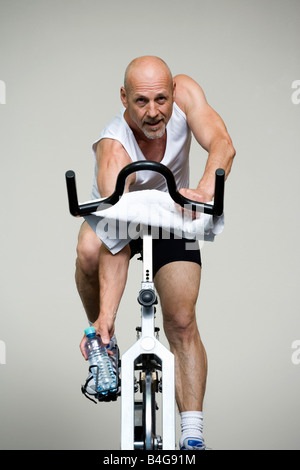 Un uomo maturo su una bicicletta stazionaria di raggiungere per una bottiglia di acqua Foto Stock