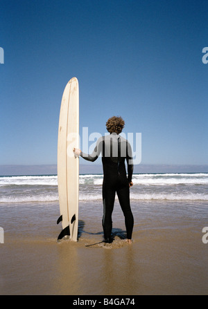 Un surfista permanente sulla spiaggia con la sua tavola da surf a guardare l'oceano Foto Stock