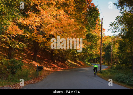 Ciclista corse su una strada rurale passato un rosso fiammante acero in autunno nel West Newbury, Massachusetts Foto Stock