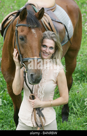 Ritratto di una bella donna bionda in piedi in un campo verde mentre tiene il suo cavallo. Foto Stock